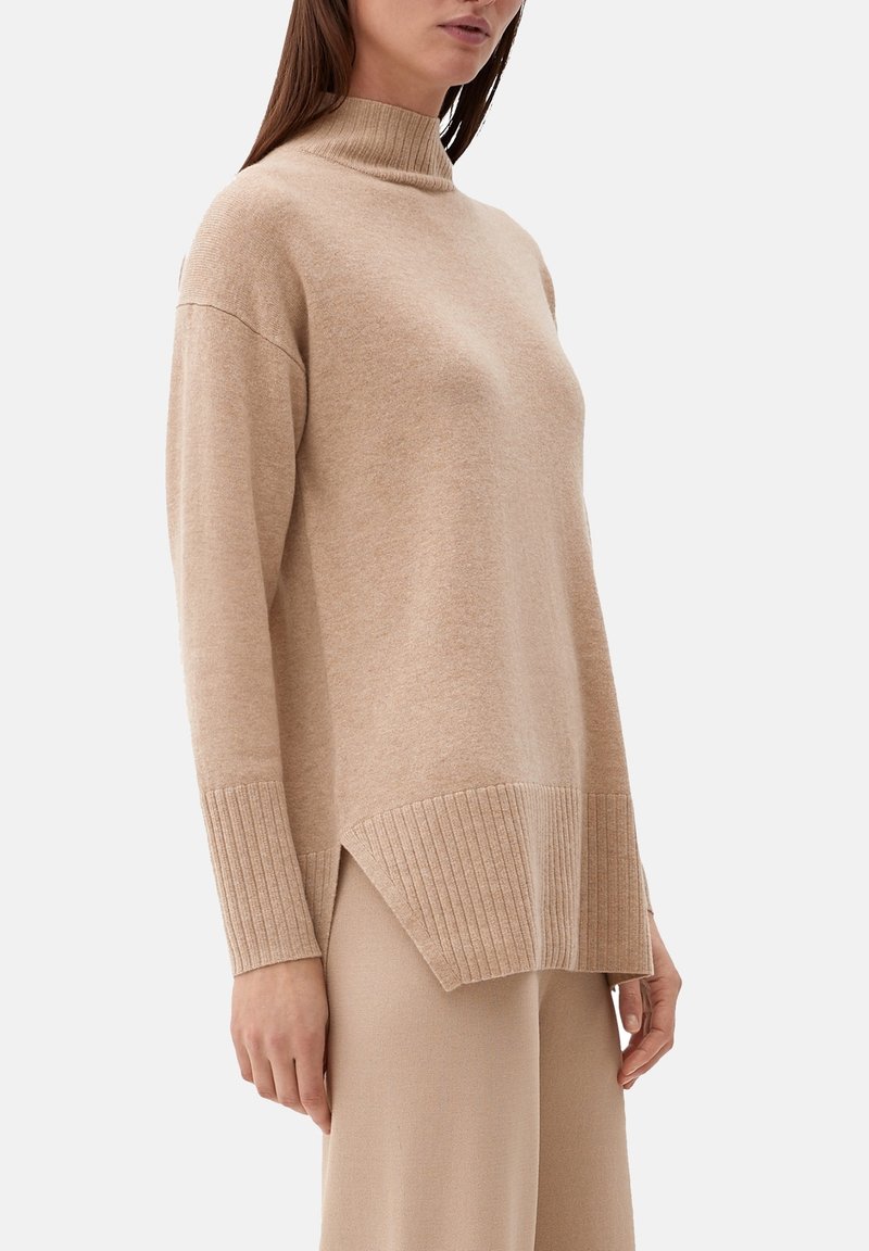 Femme portant un pull beige à col montant avec des poignets et un ourlet côtelés, associé à un pantalon beige assorti sur un fond uni.