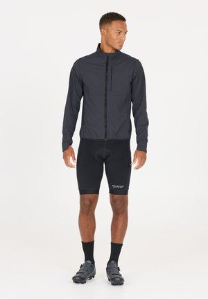 Lichtgewicht zwarte fietsjack met lange mouwen, hoge kraag en een ritszak aan de voorkant, in combinatie met zwarte fietsbroek en schoeisel.
