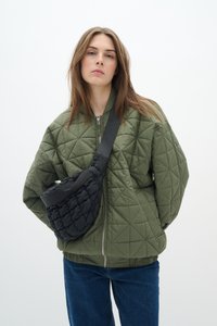 Veste bomber matelassée olive avec une fermeture éclair argentée, associée à un sac bandoulière noir texturé. Le modèle se tient devant un fond blanc.