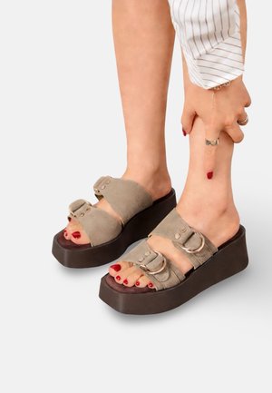 Personne portant des sandales à plateforme beige avec deux brides à boucle, du vernis à ongles rouge sur les orteils, et pointant vers sa cheville droite.