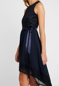 Robe marine avec un corsage en dentelle, ourlet asymétrique et ceinture en satin nouée à la taille. La dentelle texturée et le tissu lisse contrastent dans le design.