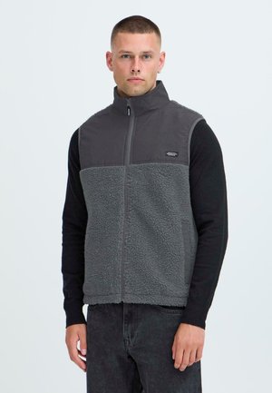 Grå fleece vest med høj krave, lynlås foran og kontraststof på brystet. Bæres over en sort langærmet skjorte med tekstureret overflade.