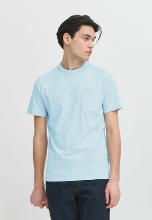 Light blue cotton t-shirt with short sleeves, rounded neckline, and a left chest pocket. Smooth texture, casual fit, paired with dark denim.