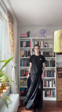 Saia preta, fluida, com uma fenda alta, combinada com uma t-shirt preta. O fundo apresenta uma estante cheia de livros e itens decorativos.