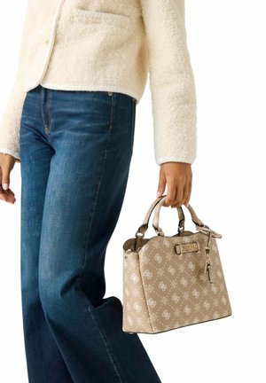 Personne portant une veste en polaire crème et un jean bleu tenant un sac à main beige Guess avec un motif blanc et des doubles poignées supérieures.