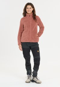Fleece-tyynytakki vaaleanpunaisena, jossa on aaltoileva rakenne, yhdistetty mustiin ja harmaisiin cargo-housuihin, joissa on sivutaskut ja vahvistetut polvet.