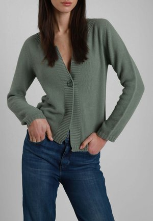 Donna che indossa un cardigan verde lavorato a maglia con una sola fibbia allacciata, abbinato a jeans blu, con le mani nelle tasche frontali su sfondo neutro.
