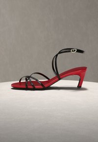 Κόκκινο σανδάλι με χαμηλό τακούνι και λεπτά μαύρα λουριά που διασχίζουν τα δάχτυλα, λουρί στον αστράγαλο με χρυσή αγκράφα, εκτεθειμένο σε ουδέτερη επιφάνεια.