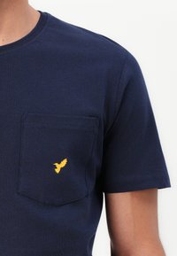 Tamno plava pamučna majica s okruglim izrezom, kratkim rukavima i ravnom džepom. Sadrži malu žutu pticu izvezenu na džepu.