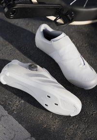 Scarpe da ciclismo bianche con una superficie liscia, un design aerodinamico e una chiusura a manopola. Le suole presentano fori per l'attacco dei pedali.