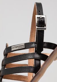 Sandal de cuero negro con múltiples tiras entrelazadas, una hebilla de plata y una suela de cuero marrón. Nombre de la marca visible en la tira.