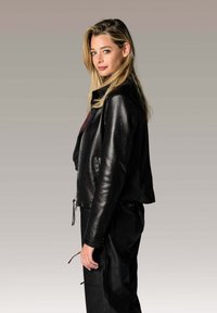 Veste en cuir noir avec une coupe ajustée, fermeture éclair et poches latérales. Le mannequin a de longs cheveux blonds et la porte avec un pantalon noir.