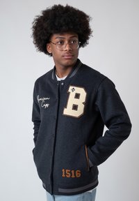 Young man with afro and glasses wearing a black varsity jacket with patches "B," "1516," and "Bavarian Caps," hands in pockets, looking right.