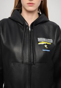 Schwarze Lederkapuzenjacke mit Reißverschluss vorne, verstellbarem Kordelzug und Fronttasche. Verfügt über ein mehrfarbiges gesticktes Logo und florales Muster.