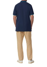 Marineblaues Poloshirt mit kurzen Ärmeln und türkisfarbenen Akzenten, kombiniert mit beigen schmalen Hosen und weißenSneakern, von hinten gesehen.