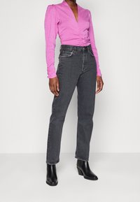 Donna con una blusa rosa a balze a maniche lunghe, jeans neri a vita alta e stivaletti neri con tacco, in piedi contro uno sfondo neutro.