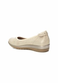 Zapato plano beige con punta redondeada, hecho de material suave, que presenta un pequeño acento decorativo en el talón y una suela flexible y texturizada.