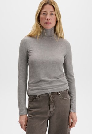 Femme aux longs cheveux blonds portant un pull à col roulé gris clair et un pantalon marron, debout sur un fond blanc uni.