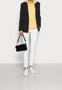 Blazer nero su un hoodie giallo, abbinato a pantaloni slim-fit bianchi e sneakers bianche. Una borsa nera aggiunge un tocco di dettaglio.