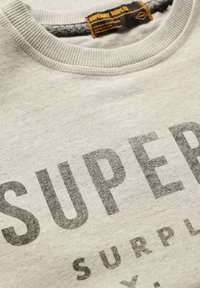 Lichtgrijs sweatshirt met geribde halslijn, voorzien van bold, donkergrijs "SUPERDRY SURPLUS"-tekst. Zachte stofstructuur, casual ontwerp.