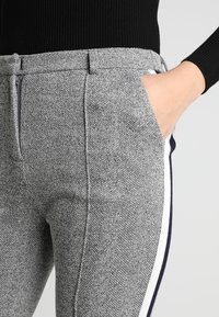 Šedé kalhoty s kostkovaným vzorem hounds tooth a přiléhavým střihem, obsahující přední kapsu a boční pruh v námořnické modré a bílé barvě. Zapínání na zip v pase.