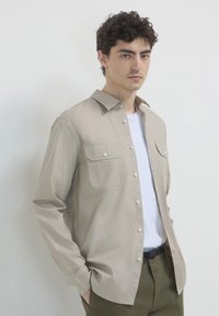 Beige Langarmhemd mit geknöpften Taschen, aus glattem Stoff gefertigt. Getragen über einem weißen T-Shirt und kombiniert mit olivgrünen Hosen.