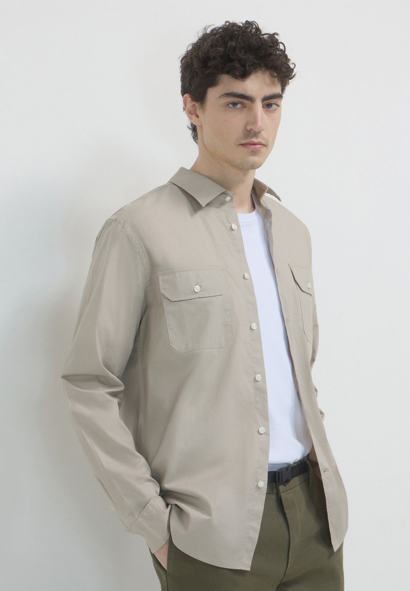Beige Langarmhemd mit geknöpften Taschen, aus glattem Stoff gefertigt. Getragen über einem weißen T-Shirt und kombiniert mit olivgrünen Hosen.