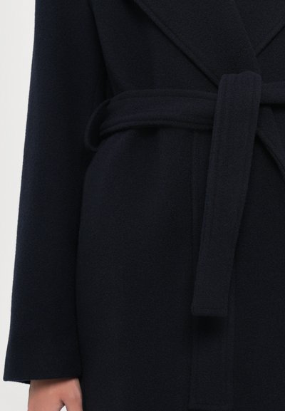 Cappotto di lana blu scuro con ampi risvolti, maniche lunghe e una cintura in tessuto abbinato annodata in vita, mostrato sul torace e sulla mano di una persona.