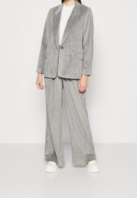 Completo in corduroy grigio con blazer a rever e pantaloni a gamba larga. Include due tasche frontali e chiusura a bottone. Abbinato a sneakers bianche.