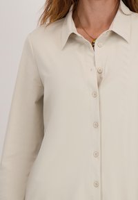 Beige overhemd met knoopsluiting, een klassieke kraag, lange mouwen en een subtiele textuur; voorzien van bijpassende knopen aan de voorkant.