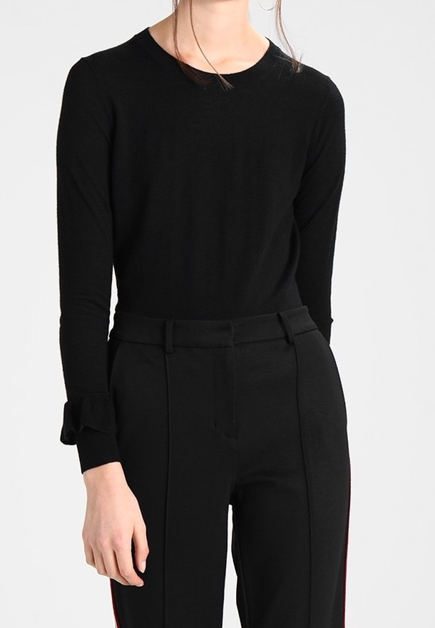Femme portant un haut noir ajusté à manches longues avec un détail nœud sur la manche et un pantalon noir taille haute avec passants pour ceinture.