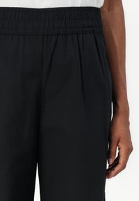 Pantaloni neri con elastico in vita, dotati di pieghe laterali, caratterizzati da una texture liscia e una vestibilità comoda. Il tessuto appare leggero e traspirante.