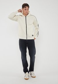 Felpa con cappuccio e zip color crema, con strisce nere sui lati, dotata di chiusura frontale, polsini a costine e un'etichetta con logo all'orlo.