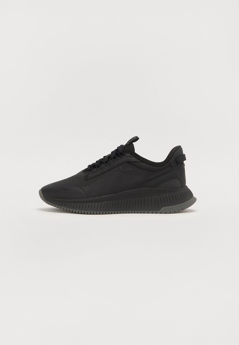 Baskets de sport noires avec dessus texturé, design à lacets, languette au talon et semelle épaisse à motifs sur un fond clair uni.