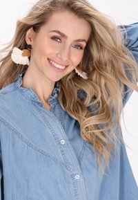 Svetlomodrá denimová košeľa s volánovým golierom a zapínaním na gombíky. Modelka má na sebe veľké, okrúhle tkané náušnice s bielymi pierkami.