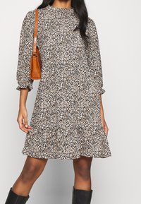 Blumenmusterkleid in Schwarz, Braun und Weiß; lange Puffärmel; rüschiger Saum; kombiniert mit einer kleinen braunen Umhängetasche; das Model trägt schwarze Stiefel.