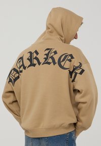 Felpa oversize beige con stampa gotica nera "BARKER" sulla schiena. Realizzata in tessuto morbido, presenta un cappuccio con cordino e un cinturino a coste.