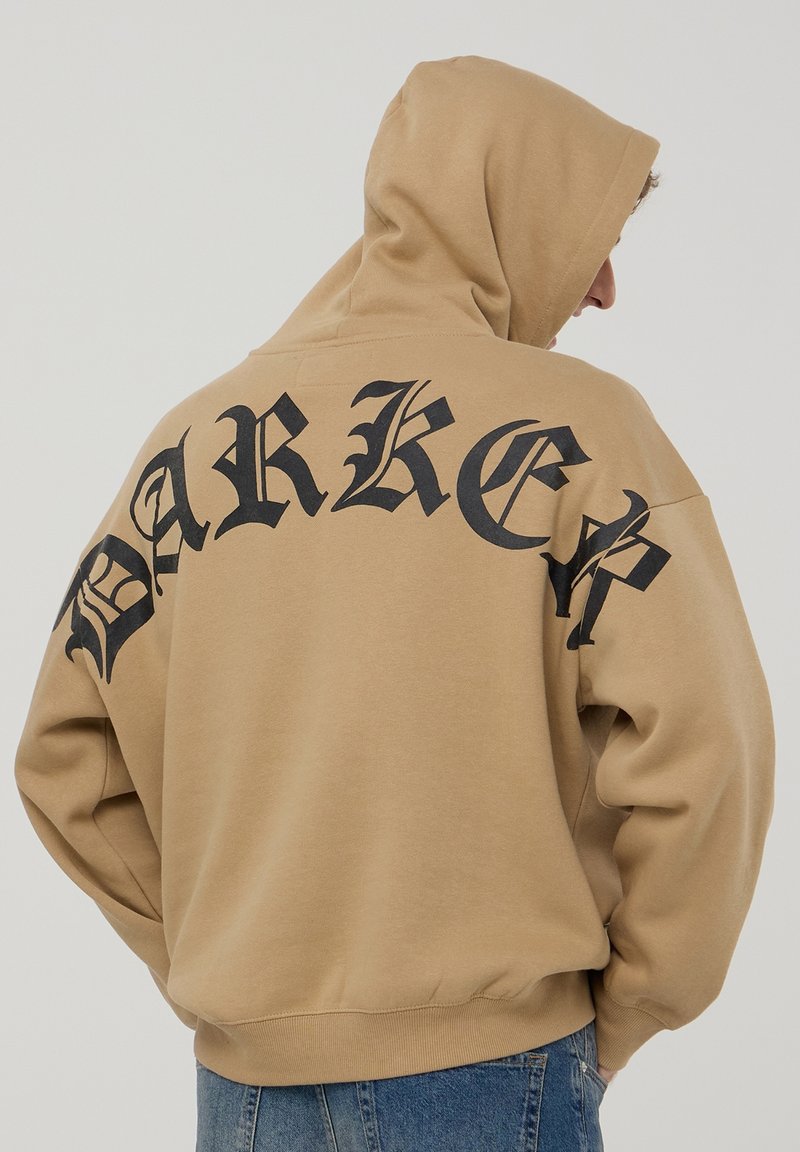 Felpa oversize beige con stampa gotica nera "BARKER" sulla schiena. Realizzata in tessuto morbido, presenta un cappuccio con cordino e un cinturino a coste.