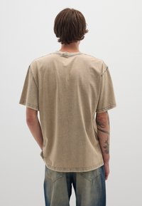 T-shirt beige a maniche corte con finitura testurizzata, dettagli di cucitura visibili e vestibilità rilassata, abbinato a jeans denim larghi di colore blu chiaro.