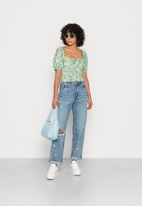Top cropped com padrão floral em verde e creme, jeans de ganga desbotados de cintura alta, bolsa azul e sapatilhas brancas.