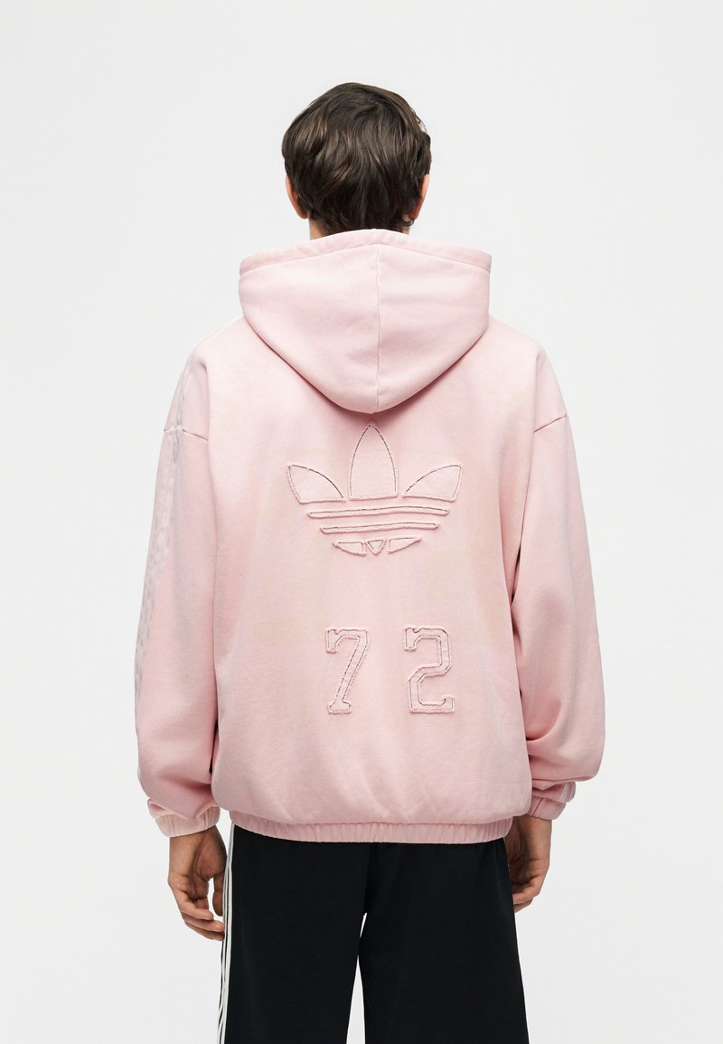 Personne portant un sweat à capuche rose clair avec un grand logo Adidas et le numéro 72 brodé dans le dos, associé à un pantalon noir avec des bandes blanches.
