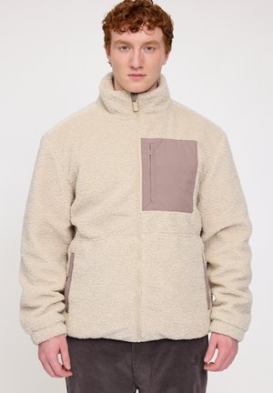 Beige Fleecejacke mit hohem Kragen und braunem Brusttasche. Mit Reißverschluss und seitlichen Taschen ausgestattet, konzipiert für Wärme und Komfort.