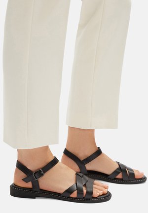 Černé kožené sandály s překříženými pásky a přezkou, s plochou podrážkou a jemným prošitím podél okraje.