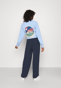 Sweatshirt bleu clair avec un motif graphique représentant des montagnes et des arbres au dos, associé à un pantalon large bleu marine et des baskets blanches.
