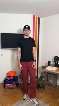 Sort t-skjorte, burgunder bukser, joggesko. Tatoveringer på armene. Svart caps med logo. Bakgrunnen inkluderer dekorativ vegg, lampe og møbler.