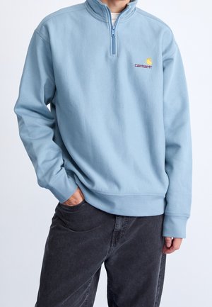 Person iført lyseblå quarter-zip sweatshirt med Carhartt-logo, sorte bukser, den ene hånd i lommen mod en ensfarvet baggrund.