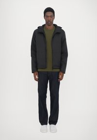 Schwarze Pufferjacke mit Kapuze, mit Reißverschluss vorne, kombiniert mit einem olivgrünen Shirt und dunkelblauen Jeans. Weiße Sneaker vervollständigen das Outfit.