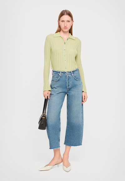 Haut à boutons côtelé vert clair avec un col, associé à un jeans bleu à jambes larges et des mules blanches à talons hauts. Sac noir à la main.