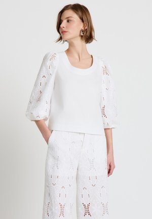 Femme portant un chemisier blanc à manches bouffantes en dentelle et un pantalon large assorti en dentelle, debout avec une main dans la poche, regardant sur le côté.