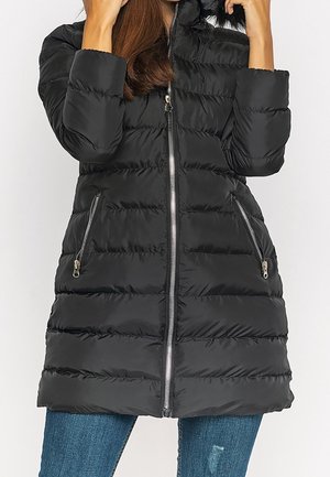 Femme portant une doudoune matelassée noire avec fermeture éclair à l'avant et poches zippées, associée à un jean bleu.